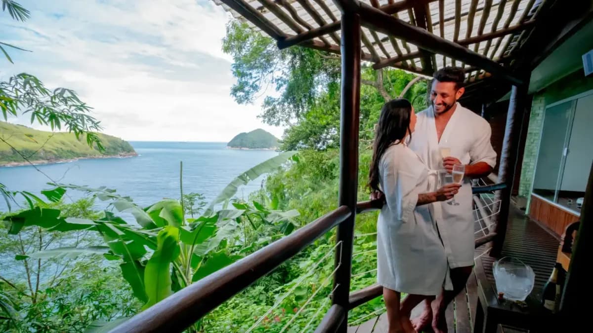 Casal de roupão brinda na varanda de madeira da suíte, com a mata e o mar ao fundo, no Ilha de Toque Toque Eco Hotel