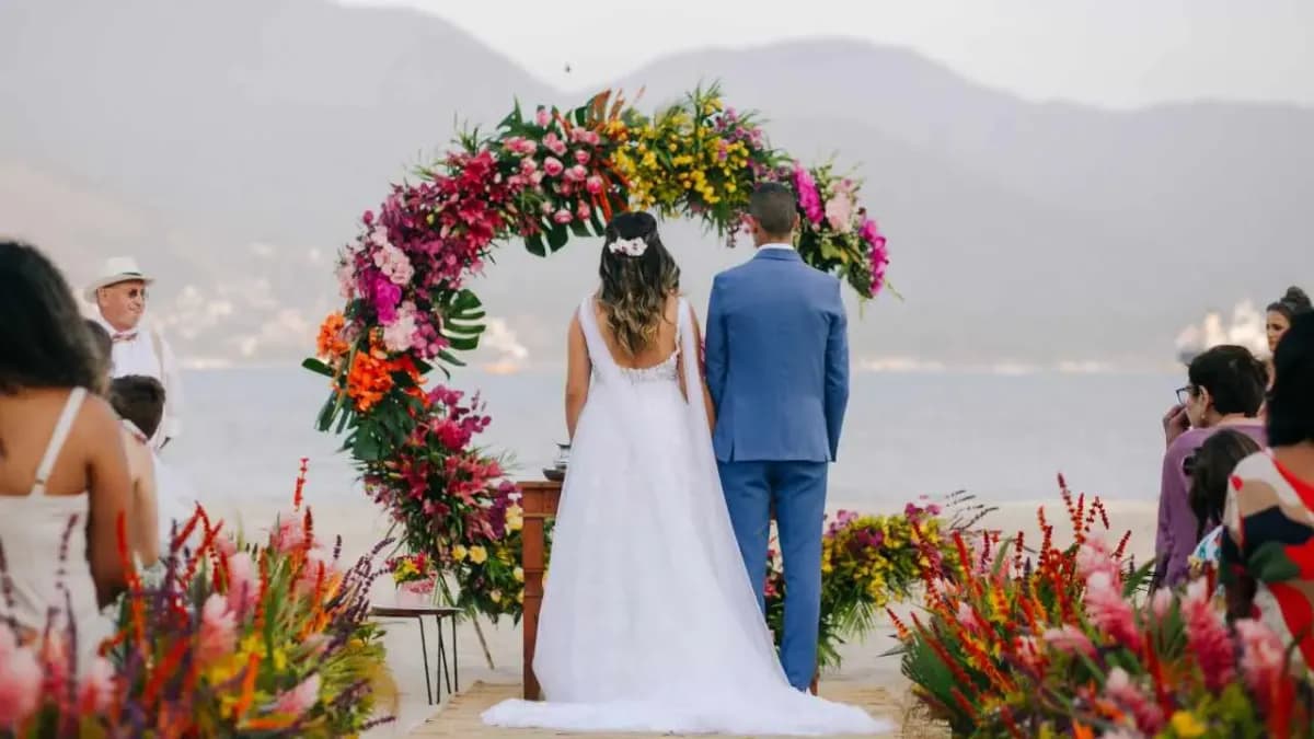 Noivos de costas diante de arco de flores coloridas em cerimônia ao ar livre, com o mar e a serra ao fundo