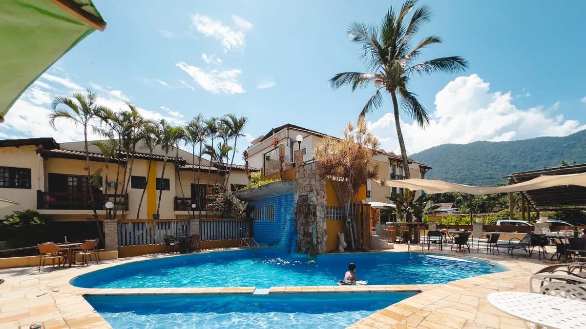 Piscina do Villa'l Mare Hotel cercada de coqueiros e prédios baixos, com a serra coberta de mata ao fundo, em Maresias