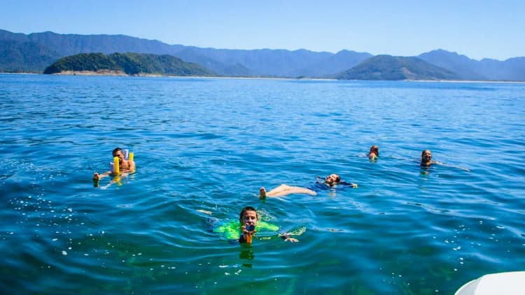 Grupo boia em mar azul e calmo com espaguetes de piscina ao lado da lancha, com ilhas e serra ao fundo, na Costa Sul de São Sebastião