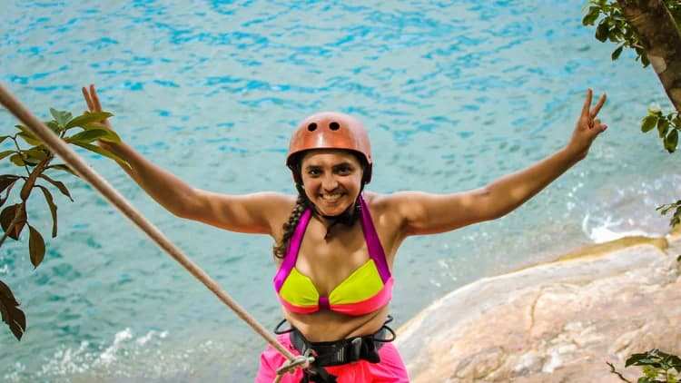 Participante de capacete sorri de braços abertos presa à corda do Rapel do Mirante, com o mar azul e as pedras logo abaixo