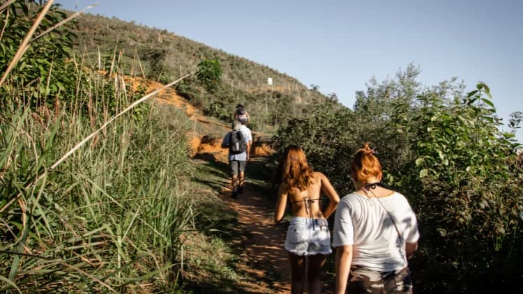 Visitantes suben un sendero de tierra entre pasto alto y arbustos hacia la cima del cerro, en Maresias, São Sebastião