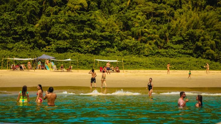 Banhistas na areia e na água verde-azulada de praia com tendas de sombra ao pé de mata densa, nas ilhas da Costa Sul de São Sebastião