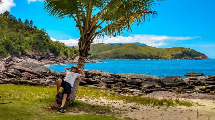 Visitante descansa apoyado en un cocotero sobre el césped, frente al peñón de piedras y el mar azul de la Praia de Calhetas