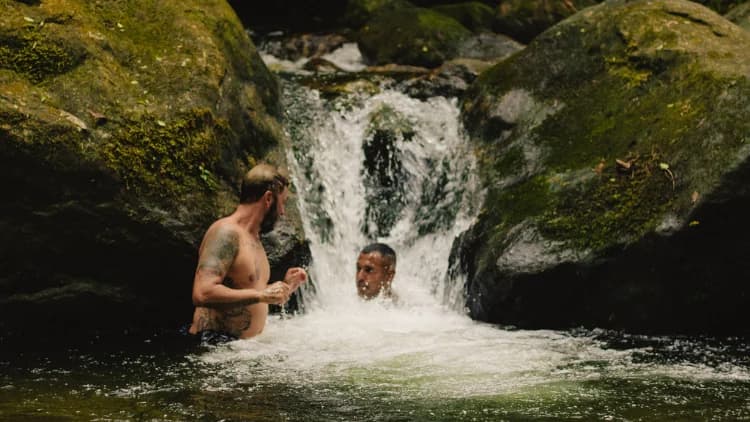 Dois visitantes se banham sob uma pequena queda d'água entre pedras com musgo, no poço de rio da mata em Maresias