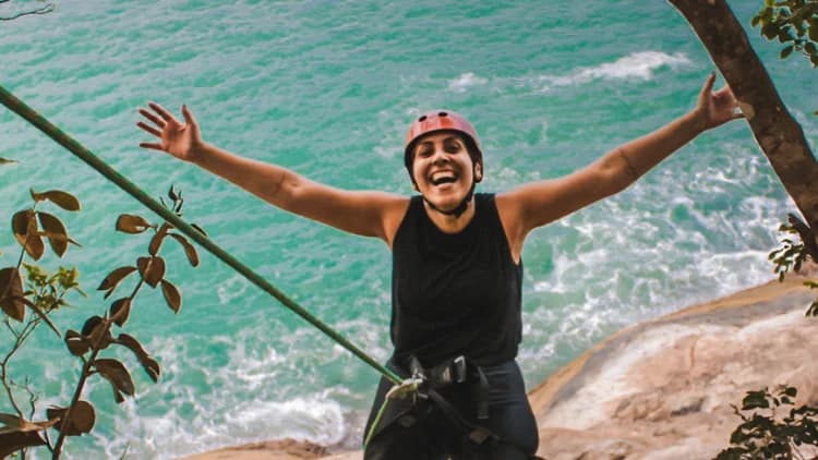 Praticante de capacete e cadeirinha abre os braços antes de descer no Rapel do Mirante, com o mar verde-azulado lá embaixo