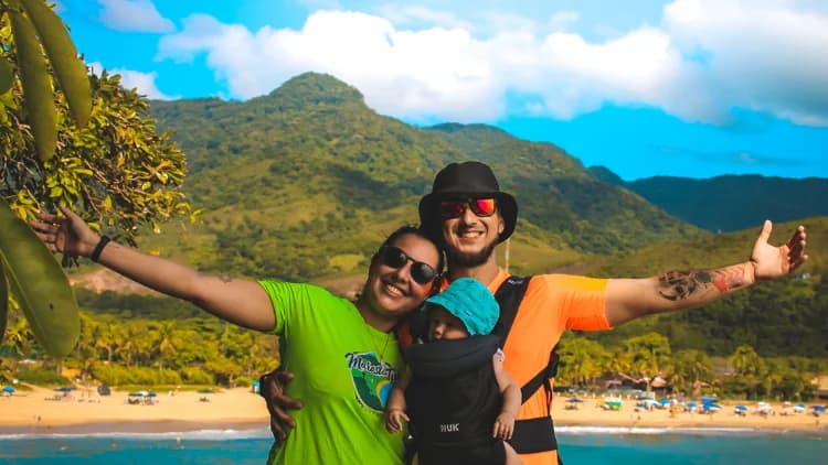 Casal com bebê no canguru abre os braços em um mirante, com a praia cheia de guarda-sóis e morros de mata ao fundo, em Maresias