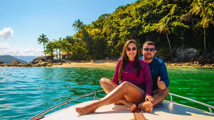 Casal sentado na proa da lancha diante de água verde, praia com coqueiros e encosta de mata, em passeio de lancha pelo litoral