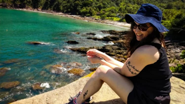 Visitante de chapéu sentada em uma pedra do costão, com a água verde-azulada e as pedras da Praia de Calhetas logo abaixo