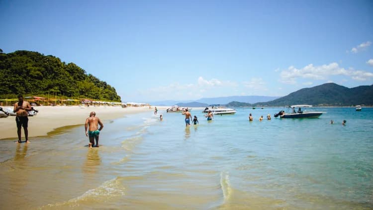 Turistas caminham na água rasa em direção a lanchas ancoradas junto a praia de areia clara e mata, nas ilhas da Costa Sul de São Sebastião