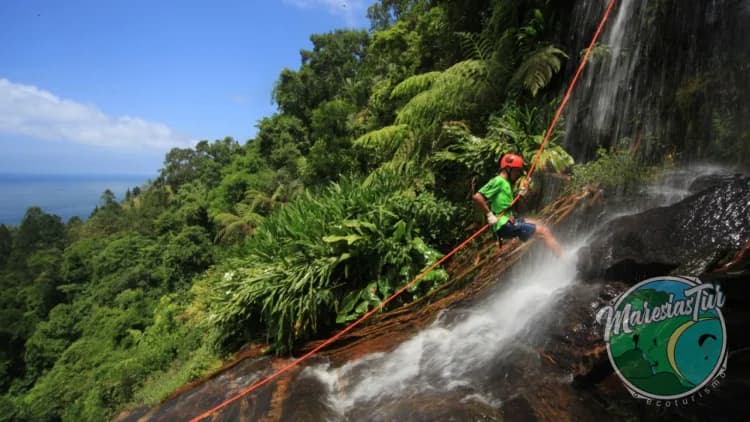 Praticante de rapel desce pela rocha molhada ao lado da queda-d'água, entre a mata, com o mar no horizonte, na Cachoeira de Calhetas