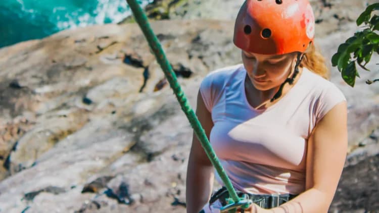 Participante de capacete segura a corda presa à cadeirinha no início da descida do Rapel do Mirante, com a rocha e o mar atrás