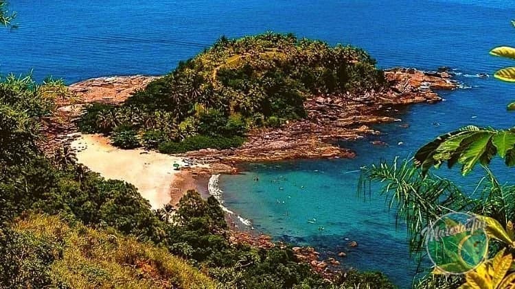 Vista do alto da enseada da Praia de Calhetas, com areia clara, água verde-azulada, costão rochoso e morro de mata em São Sebastião