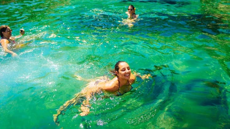 Amigas nadam sorrindo em água verde e transparente, com o fundo visível, numa parada para banho nas ilhas da Costa Sul de São Sebastião