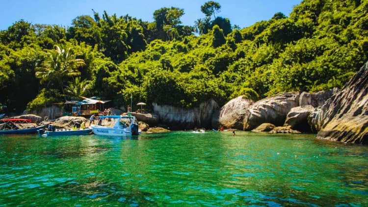 Barcos de passeio atracados em água verde junto a costão de pedras e mata densa, com banhistas, nas ilhas da Costa Sul de São Sebastião