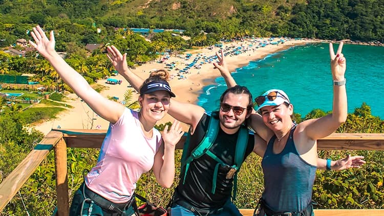 A group of three people with rappelling gear poses on the lookout deck, with the beach and ocean in the background, between Paúba and Maresias