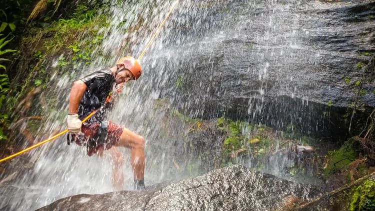 Praticante de rapel de capacete e luvas desce sob o jato d'água, apoiado na rocha, na Cachoeira de Calhetas, em São Sebastião