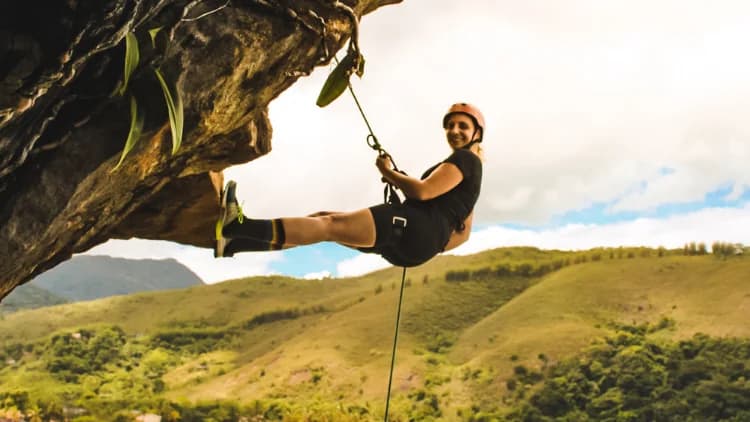 Praticante de capacete fica suspensa na corda com os pés na borda da rocha no Rapel do Mirante, com morros verdes ao fundo