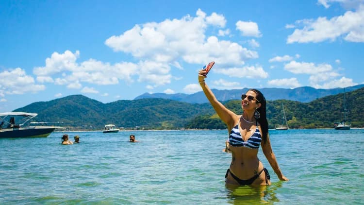 Turista em pé na água rasa e clara tira selfie, com lanchas ancoradas e montanhas cobertas de mata ao fundo, numa parada do passeio pela Costa Sul