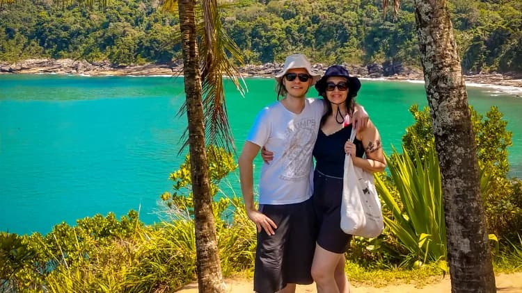 Casal abraçado posa entre coqueiros no alto da trilha, com a água turquesa da enseada da Praia de Calhetas e a mata ao fundo