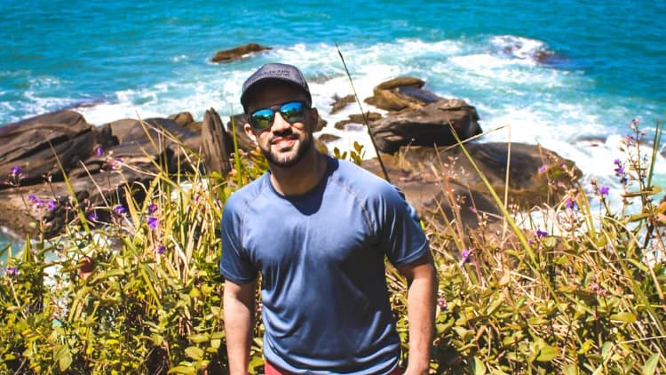 Visitante de boné posa entre capim e flores roxas, com as pedras do costão e o mar com ondas da Praia de Calhetas ao fundo
