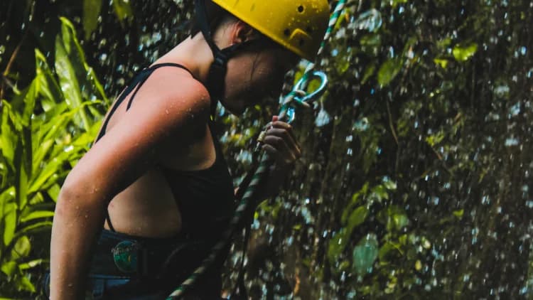 Praticante de rapel de capacete amarelo segura a corda e o mosquetão sob gotas d'água, na Cachoeira de Calhetas, em São Sebastião