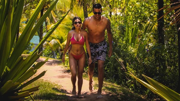 Casal em roupa de banho caminha por trilha de terra entre palmeiras e vegetação de restinga a caminho da Praia de Calhetas