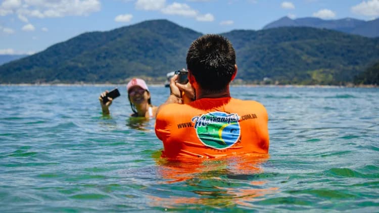 Pessoa de camiseta laranja com água na cintura fotografa turista no mar calmo, com montanhas de mata ao fundo, no passeio pela Costa Sul