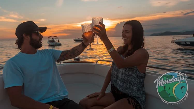 Couple toasts with glasses on the boat during sunset, with other boats anchored at sea, on the tour through the South Coast of São Sebastião