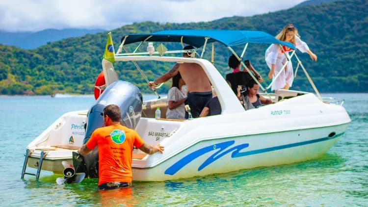 Lancha Runner 7500 branca com faixa azul e bandeira do Brasil ancorada em água rasa e verde, com grupo a bordo, no roteiro de lancha até Ilhabela