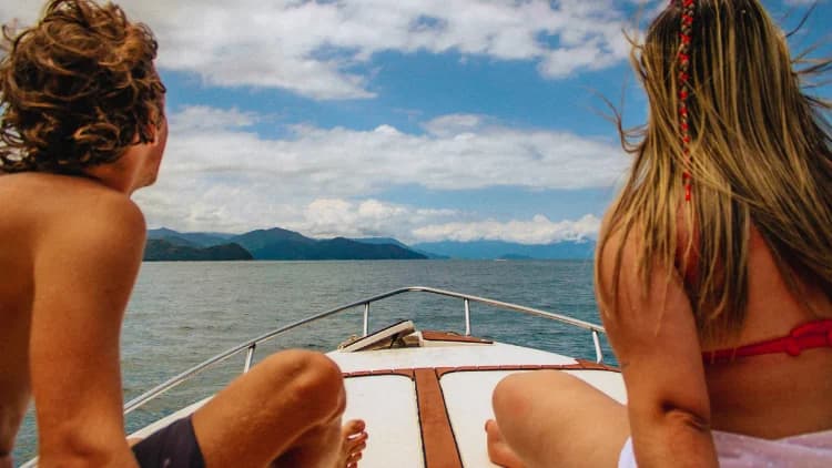 Duas pessoas de costas sentadas na proa da lancha observam o mar e as montanhas cobertas de mata, no roteiro de lancha até Ilhabela