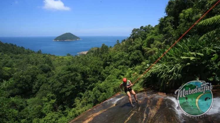 A rappeller begins the descent at the top of the Calhetas Waterfall, with the Atlantic Forest, ocean, and an island in the background