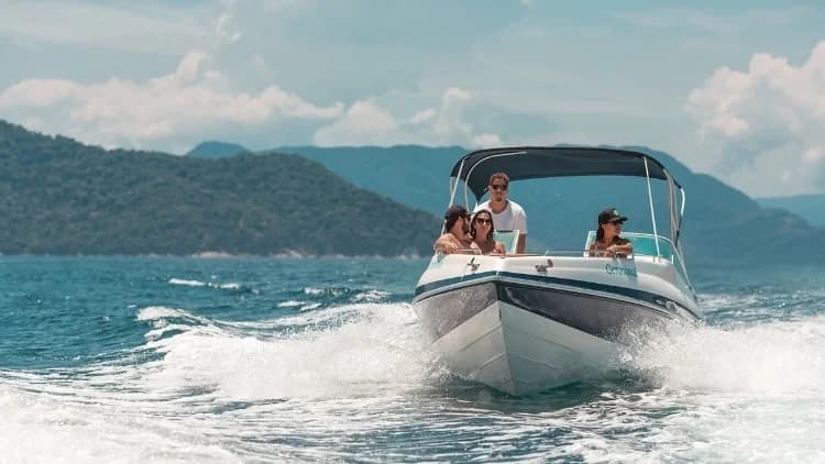 Lancha com cinco passageiros navega levantando espuma no mar, com morros cobertos de mata ao fundo, a caminho das ilhas da Costa Sul de São Sebastião
