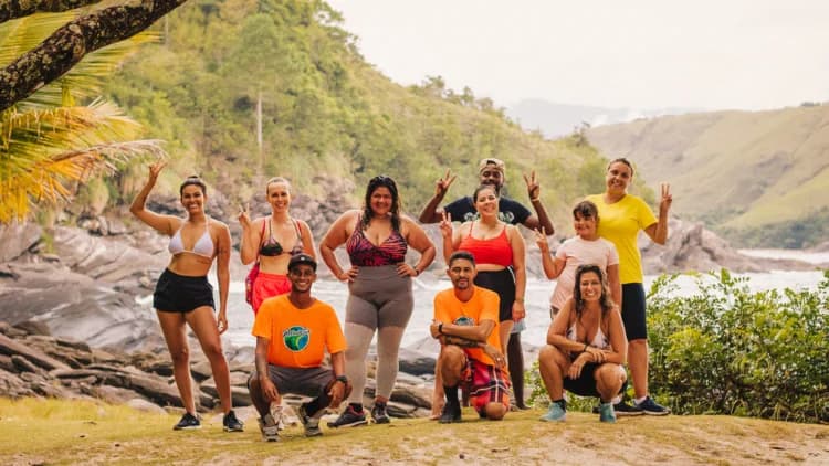 Grupo de visitantes e dois guias de camiseta laranja posam na grama, com pedras escuras, água e morros verdes ao fundo em São Sebastião