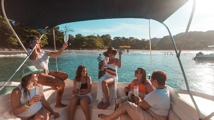 Grupo de amigas brinda com taças sob o toldo de lancha ancorada perto de praia com mata, no passeio pelas ilhas da Costa Sul de São Sebastião