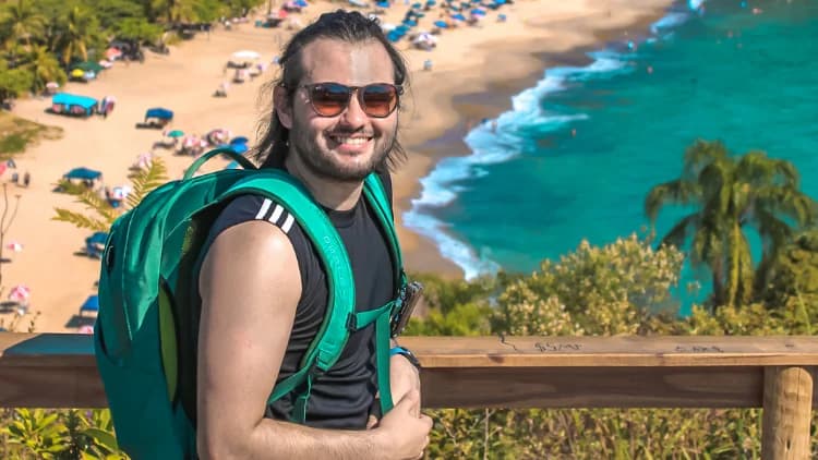 Visitante de mochila sorri no corrimão de madeira de um mirante, com a praia de guarda-sóis e o mar verde-azulado abaixo, em Maresias