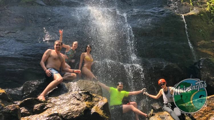 Grupo de seis pessoas posa nas pedras na base da queda-d'água da Cachoeira de Calhetas, em São Sebastião, uma delas de capacete e cadeirinha