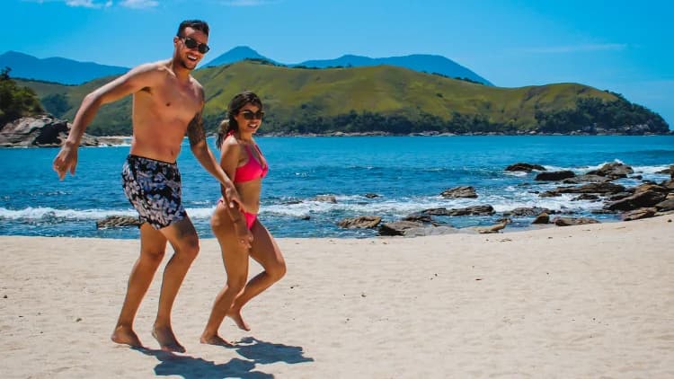 Casal em roupa de banho caminha de mãos dadas na areia da Praia de Calhetas, com mar azul, pedras e morros verdes ao fundo